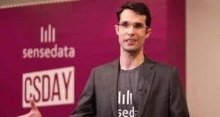 Homem de óculos e camiseta preta fala em evento diante de painel rosa com o logotipo SenseData