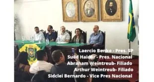 Homens sentados a uma mesa em salão com retratos antigos na parede e legenda com nomes sobreposta