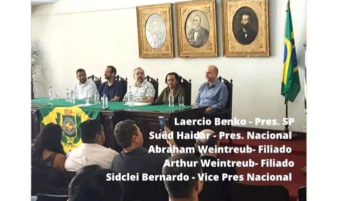Homens sentados a uma mesa em salão com retratos antigos na parede e legenda com nomes sobreposta