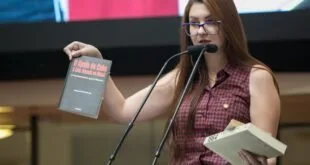 Mulher fala ao microfone em tribuna segurando dois livros, com bandeiras ao fundo