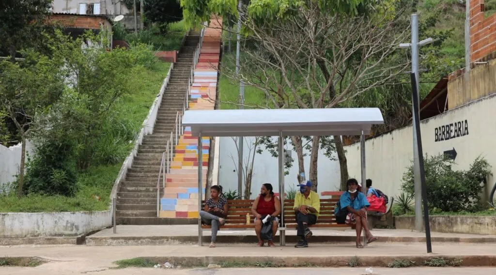 Pessoas sentadas em ponto de ônibus colorido ao lado de escadaria com árvores