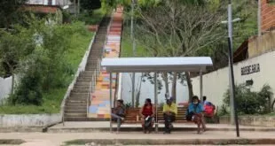 Pessoas sentadas em ponto de ônibus colorido ao lado de escadaria com árvores