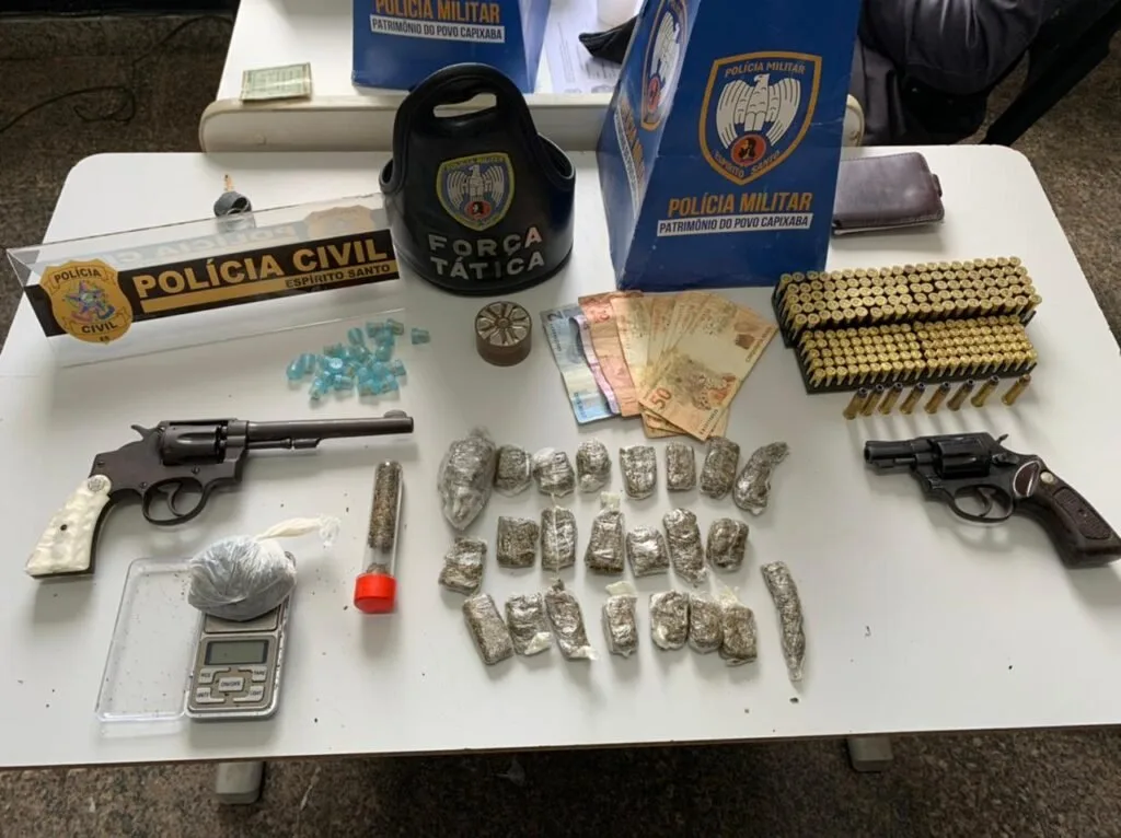 Revólver, munições e porções de droga sobre mesa ao lado de coletes da Polícia Militar