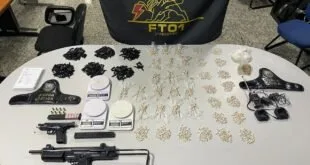 Submetralhadora, pistolas, munições e porções de droga sobre mesa diante de bandeira de batalhão