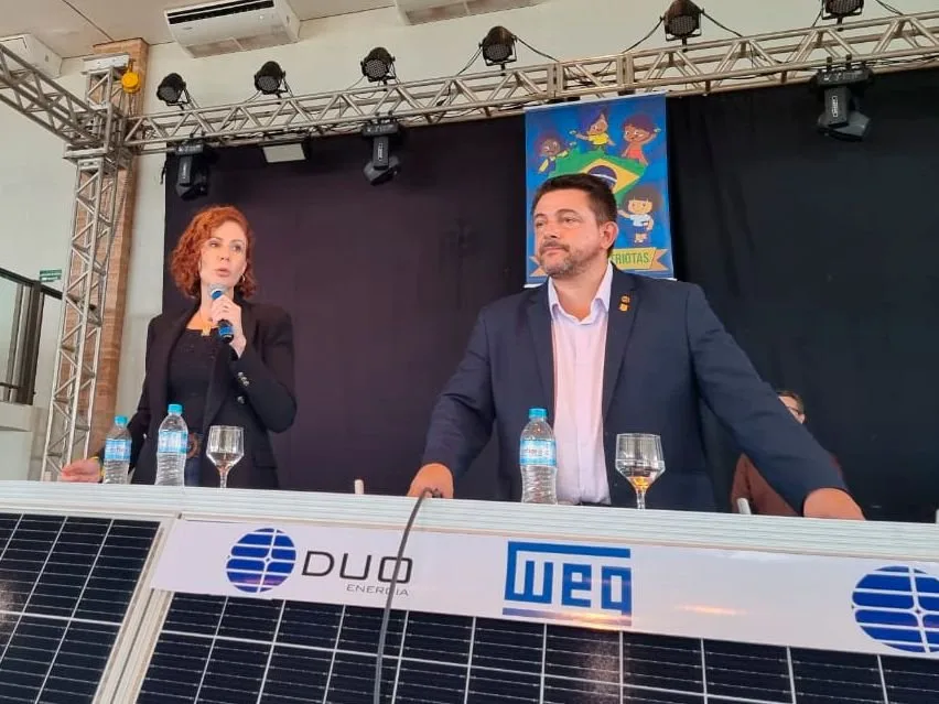 Mulher ao microfone e homem sentados a mesa com placa solar e os logotipos 'DUO' e 'WEG'