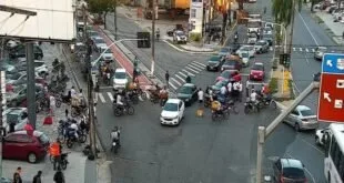 Vista do alto de cruzamento movimentado com carros e motos disputando espaço na via