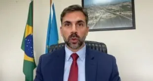 Homem de terno azul e gravata vermelha fala sentado entre a bandeira do Brasil e foto aérea de cidade