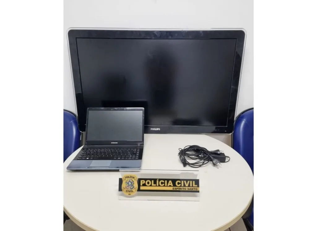 Notebook e monitor sobre mesa branca ao lado de placa com a inscrição 'Polícia Civil'