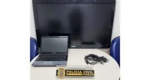 Notebook e monitor sobre mesa branca ao lado de placa com a inscrição 'Polícia Civil'