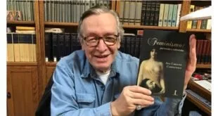 Homem idoso de óculos sorri segurando um livro diante de estante repleta de volumes