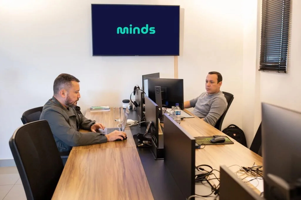 Dois homens trabalham em computadores em escritório com o logotipo 'minds' na tela da parede