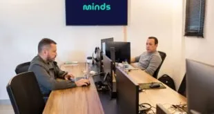 Dois homens trabalham em computadores em escritório com o logotipo 'minds' na tela da parede