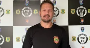 Homem de camiseta preta posa diante de painel com brasões da Polícia Federal