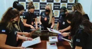 Policiais civis fardados e mascarados reunidos em torno de mesa com documentos e pastas