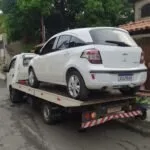 Carro branco carregado na plataforma de um guincho em rua de bairro arborizado