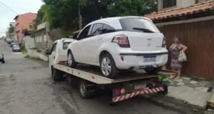 Carro branco carregado na plataforma de um guincho em rua de bairro arborizado