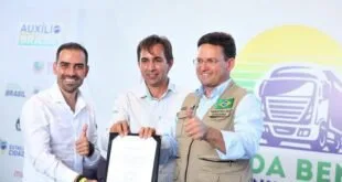 Homens sorriem exibindo documento assinado em palco durante cerimônia com painel colorido