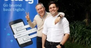 Dois homens sorriem e gesticulam diante de painel azul com o logotipo 'Lang' e a frase 'beyond English'