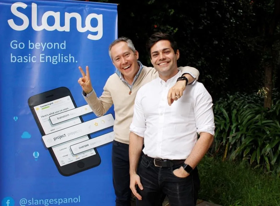 Dois homens sorriem e gesticulam diante de painel azul com o logotipo 'Lang' e a frase 'beyond English'
