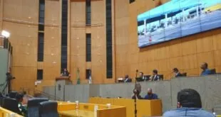 Plenário de assembleia legislativa com parlamentares nas bancadas de madeira e telões acesos