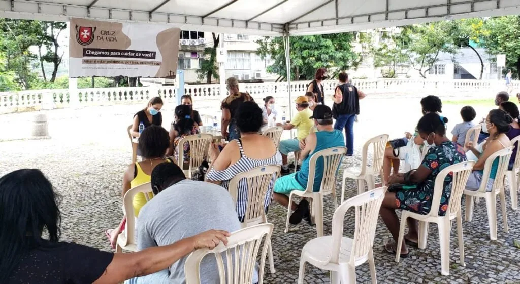 Pessoas de máscara sentadas em cadeiras de plástico aguardam atendimento em área coberta