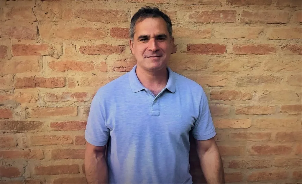 Homem de camisa polo azul sorri diante de parede de tijolos aparentes
