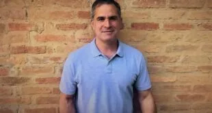 Homem de camisa polo azul sorri diante de parede de tijolos aparentes