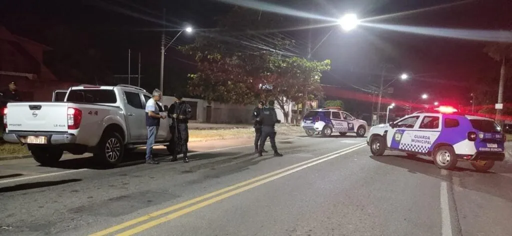 Policiais abordam pessoas em via à noite, com viatura de giroflex ligado ao fundo