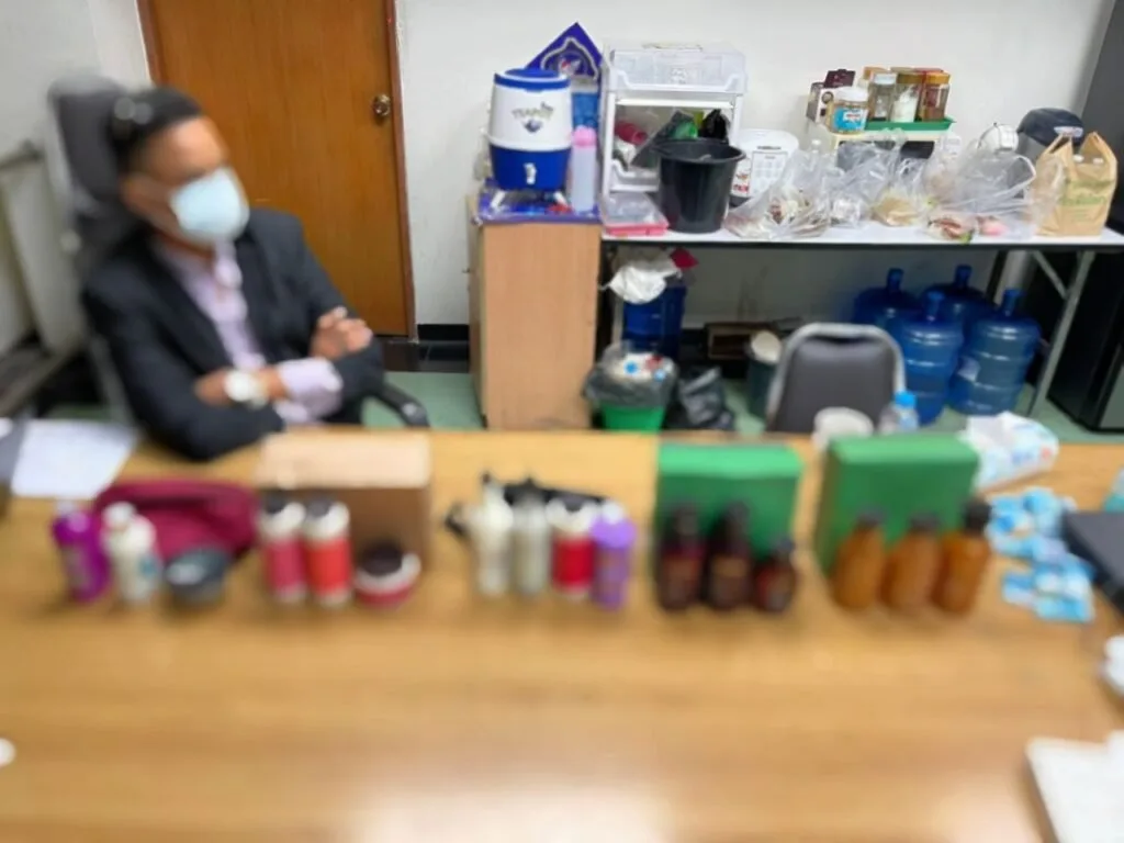 Frascos e embalagens de produtos enfileirados sobre mesa, com homem de máscara ao fundo