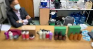 Frascos e embalagens de produtos enfileirados sobre mesa, com homem de máscara ao fundo