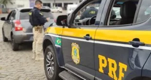Agente da PRF em pé ao lado de viatura com o brasão do órgão na lateral