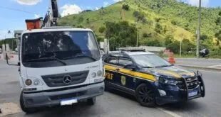 Caminhão branco e viatura da PRF estacionados lado a lado em estrada com morros ao fundo