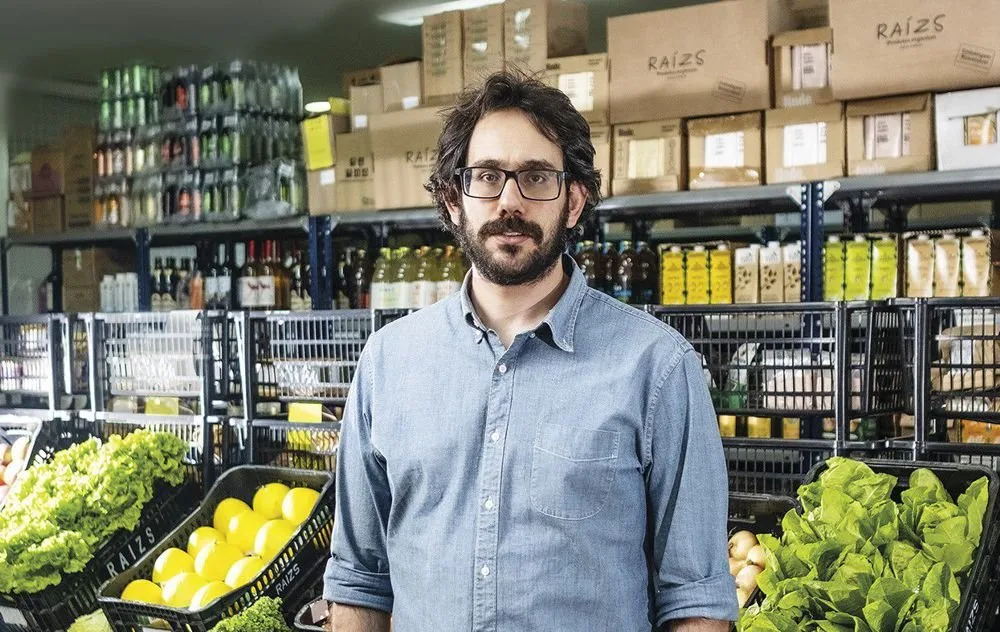Homem de óculos posa em corredor de mercado entre prateleiras de alimentos e frutas