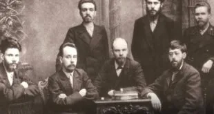 Retrato antigo em preto e branco de quatro homens de terno posando ao redor de uma mesa
