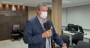 Homem de terno e máscara de proteção fala ao microfone durante entrevista em sala institucional