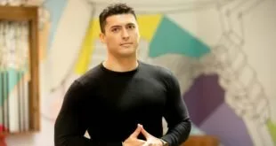 Homem jovem de blusa preta posando em ambiente interno com painel colorido ao fundo