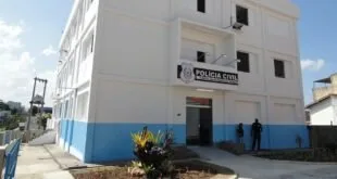 Fachada de prédio branco e azul com placa da Polícia Civil e agentes junto à entrada