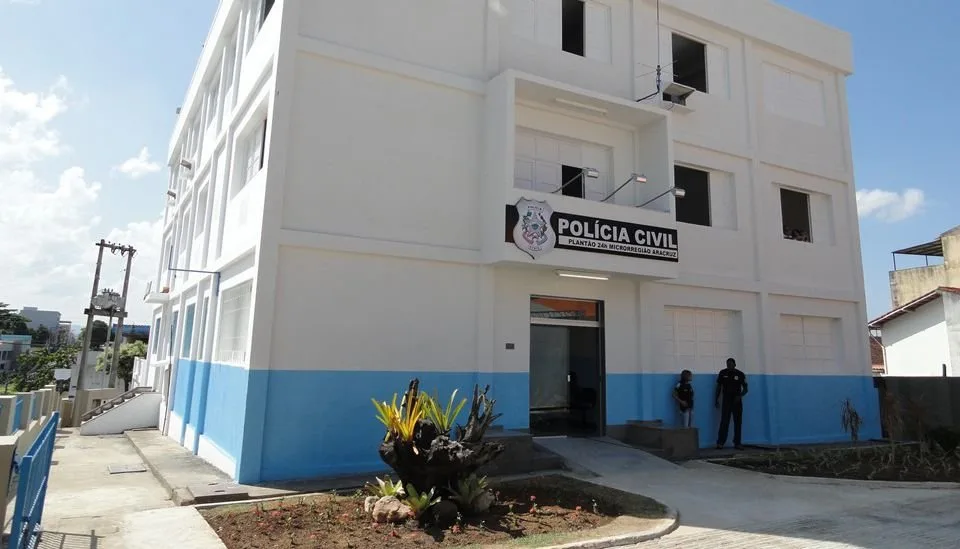 Fachada de prédio branco e azul com placa da Polícia Civil e agentes junto à entrada