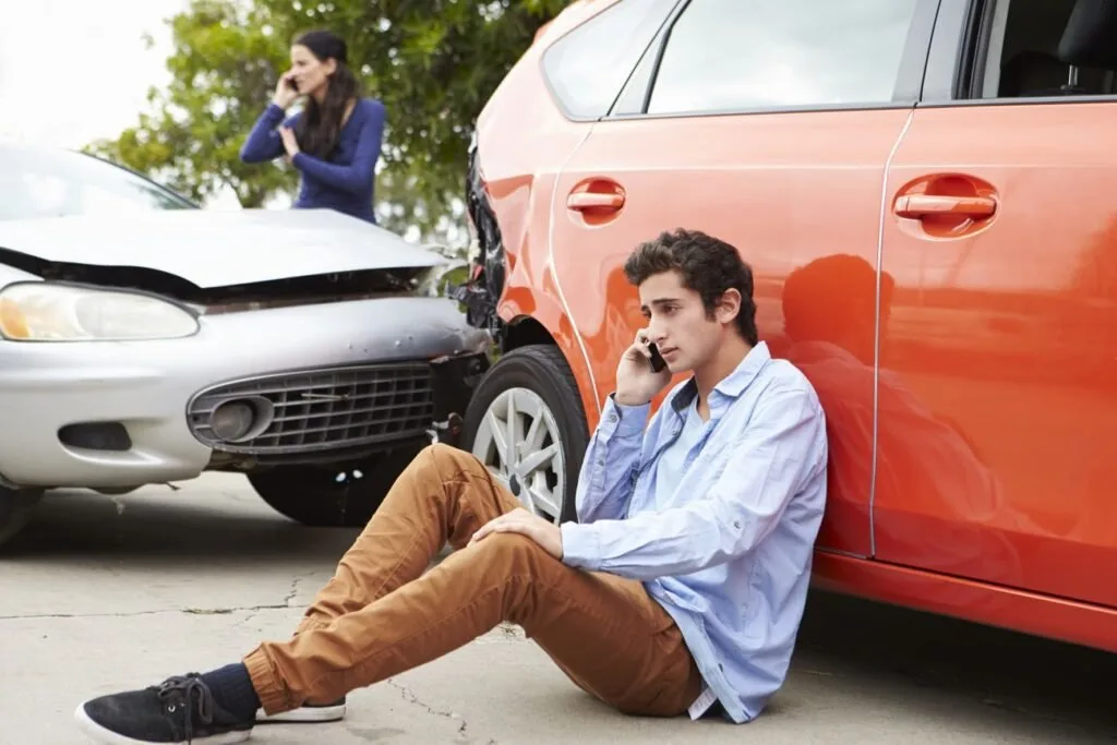 Homem sentado no asfalto ao lado de carro vermelho amassado fala ao celular após colisão