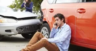 Homem sentado no asfalto ao lado de carro vermelho amassado fala ao celular após colisão