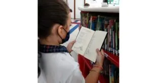Estudante de uniforme e máscara lê um livro em frente a estantes de biblioteca