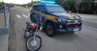 Motocicleta vermelha ao lado de viatura da PRF com giroflex aceso em via asfaltada