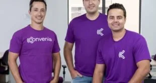 Três homens de camiseta roxa da mesma empresa posam juntos em escritório