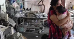 Mulher com criança no colo em oficina improvisada cercada de sucata e ferramentas