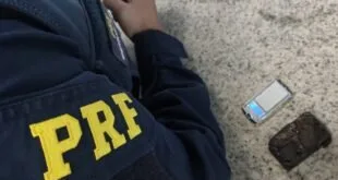Braço de agente com jaqueta escrita PRF ao lado de celular caído no chão