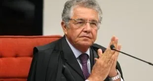 Homem idoso de terno gesticula com a mão enquanto fala sentado em bancada