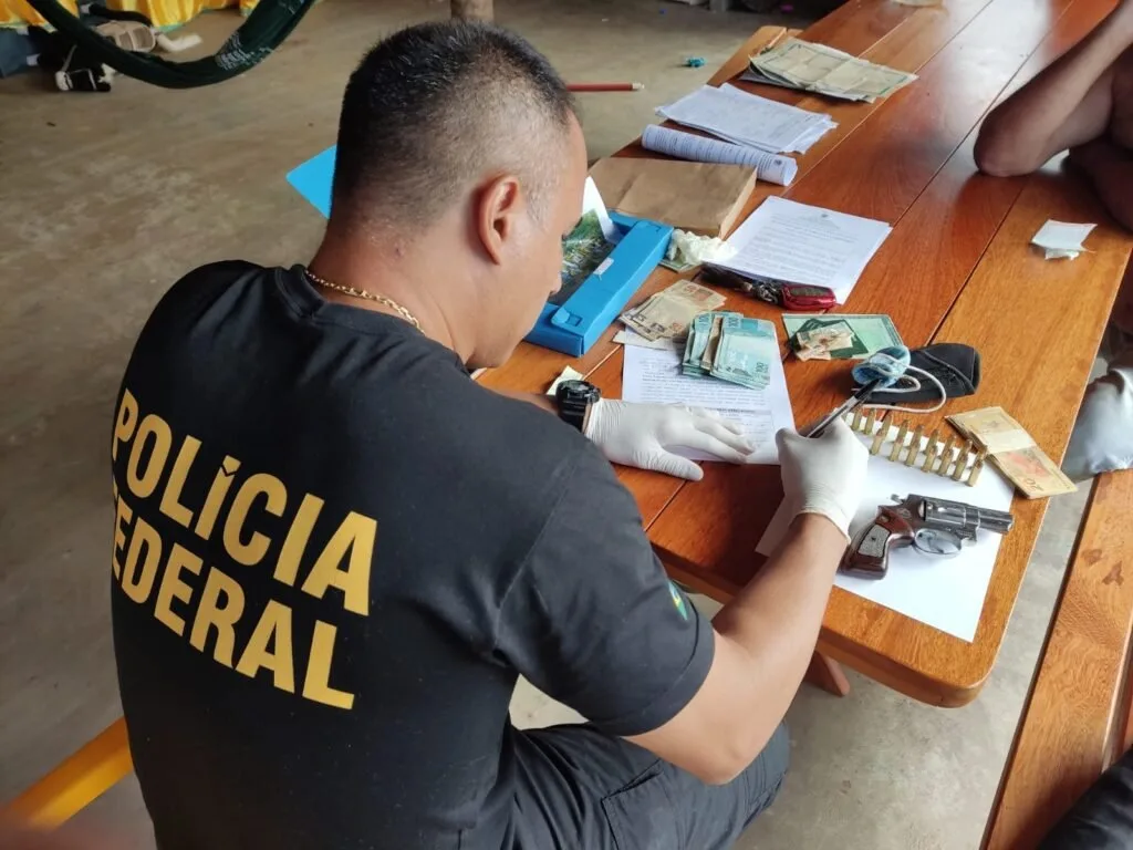 Agente com camiseta da Polícia Federal cataloga cédulas, revólver e munições sobre mesa de madeira