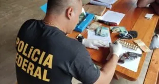 Agente com camiseta da Polícia Federal cataloga cédulas, revólver e munições sobre mesa de madeira