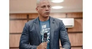 Homem de blazer fala ao microfone diante de púlpito em ambiente com paredes de madeira