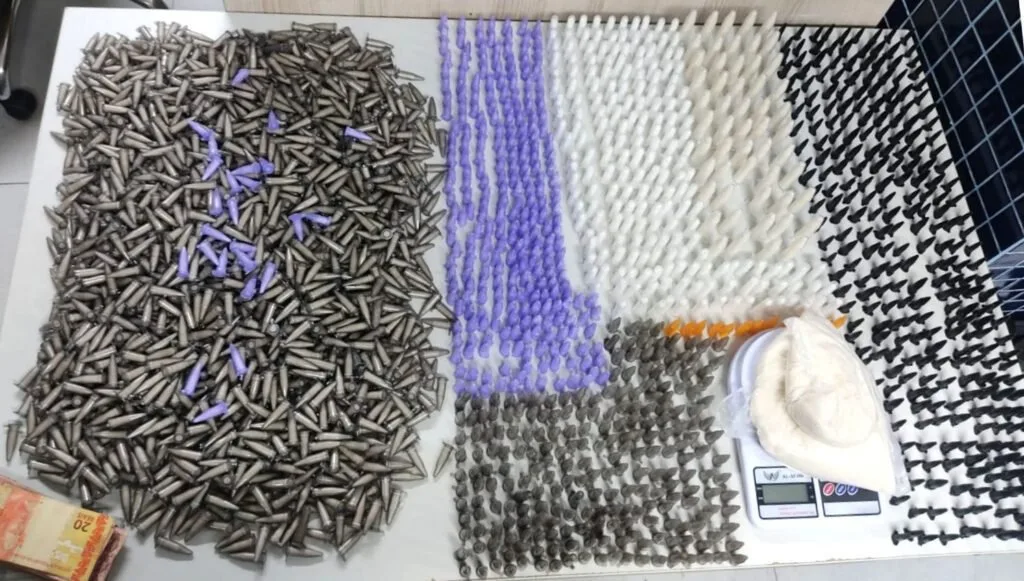 Milhares de pinos e trouxinhas de droga alinhados sobre mesa ao lado de balança e munições